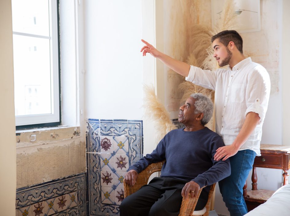 Un jeune homme accompagne un homme âgé dans un intérieur lumineux, en lui montrant la fenêtre